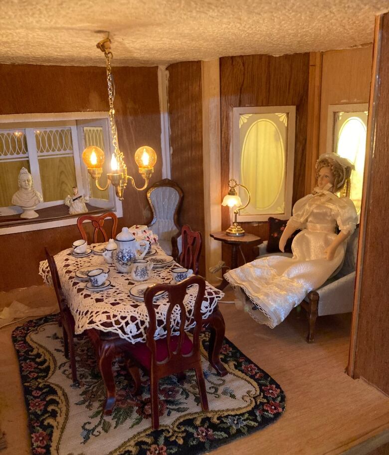 A doll sitting on a miniature couch on the right. A miniature dining room table on the left on top of a rug, with a chandelier hanging above it.