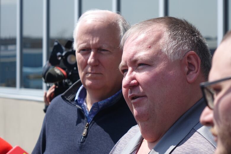 Two men speak to a group of people outside of an office building. 