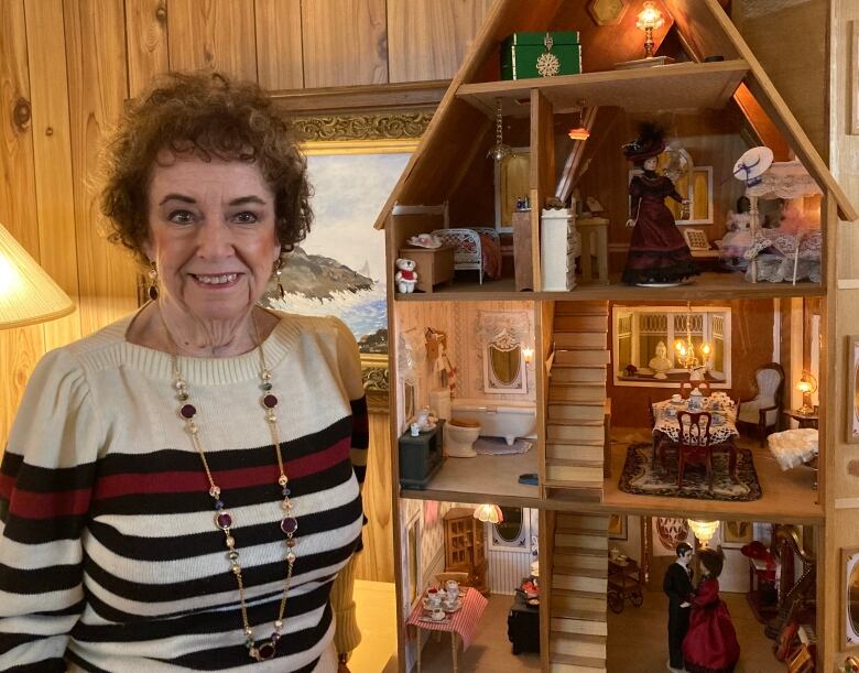 A woman, left, standing next to a dollhouse that is taller than her. Seven open-face rooms are filled with tiny pieces of furniture.