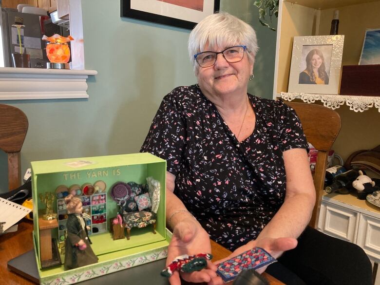 A woman, right, holding tiny knit projects. On the left, a box holding a doll and miniature furniture.