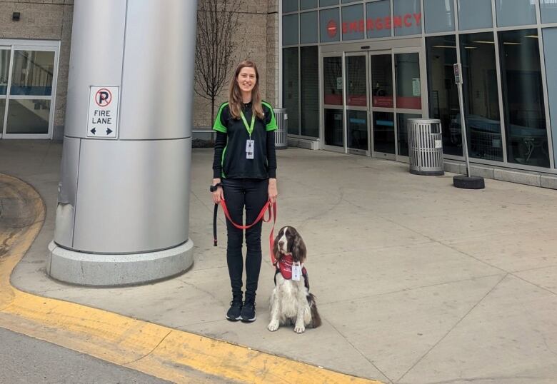 A young woman standing outside a hospital with a dog on a leash. 