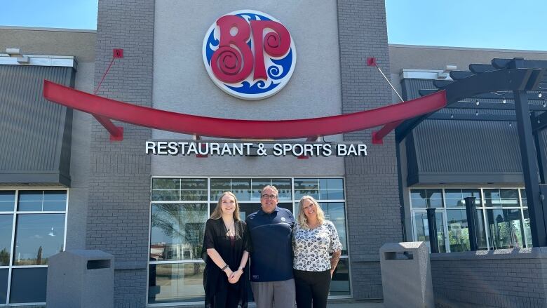 Three people stand outside a Boston Pizza restaurant on a sunny day.