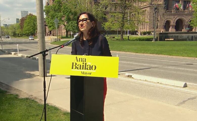 Photo of Ana Bailo at a podium with the Ontario Legislature in the background. 