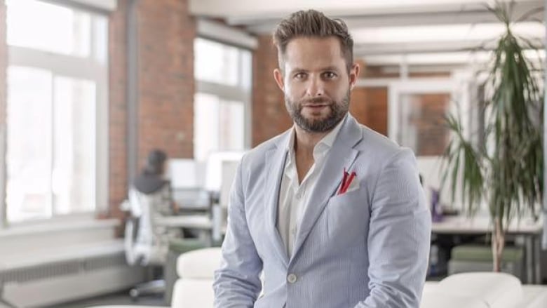 A young bearded man in a light suit and an open-necked white shirt sits on the edge of a white couch in a tastefully decorated apartment, looking directly into the camera with a neutral expression.