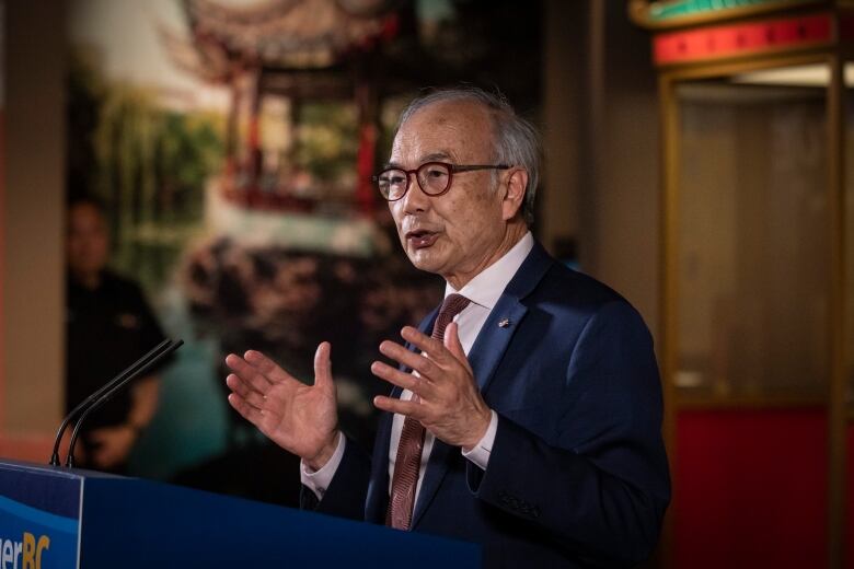 A man with glasses stands making an announcement at a podium. 