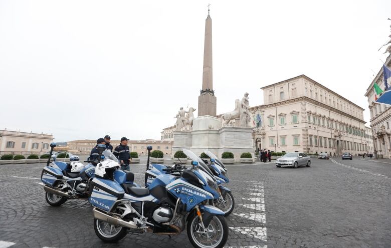 Four unmanned police motorcyles are parked by European-style buildings.