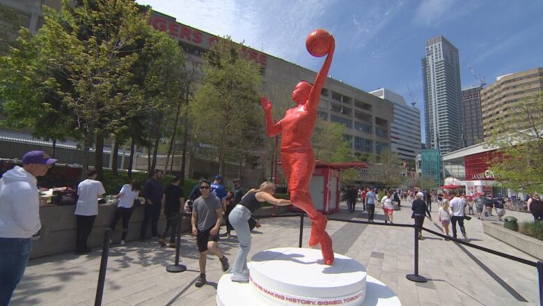 A red sculpture is seen outside, where people are passing by and interacting with it.