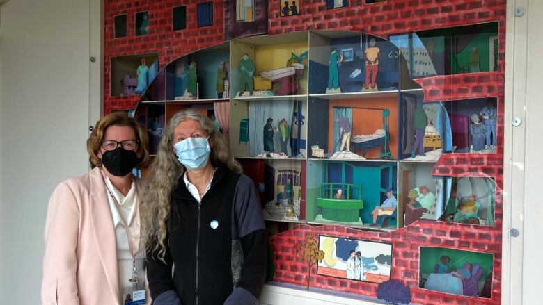 Two women wearing face masks stand in front of an art display, looking toward the camera.