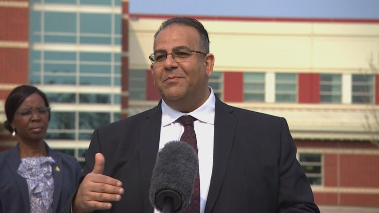 A surgeon in a dark blazer, white shirt and reddish tie speaks into a microphone at a news conference.