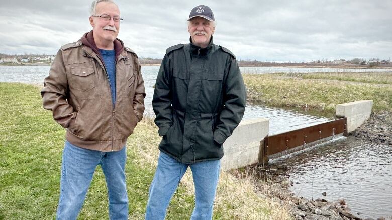 Two man stand on grass in front if a small dam