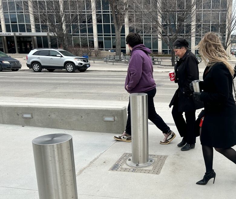 A woman wearing black walks on a sidewalk with another woman wearing a black jacket and a person wearing a purple hoodie.