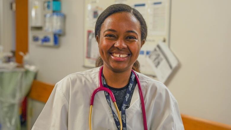 A woman wearing white scrubs and a pink stethoscope poses in a hospital.