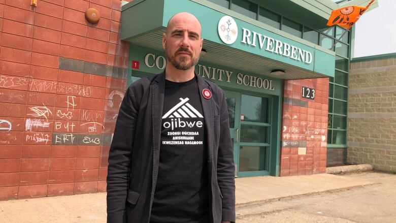 A man in a black t-shirt stands in front of a school. 