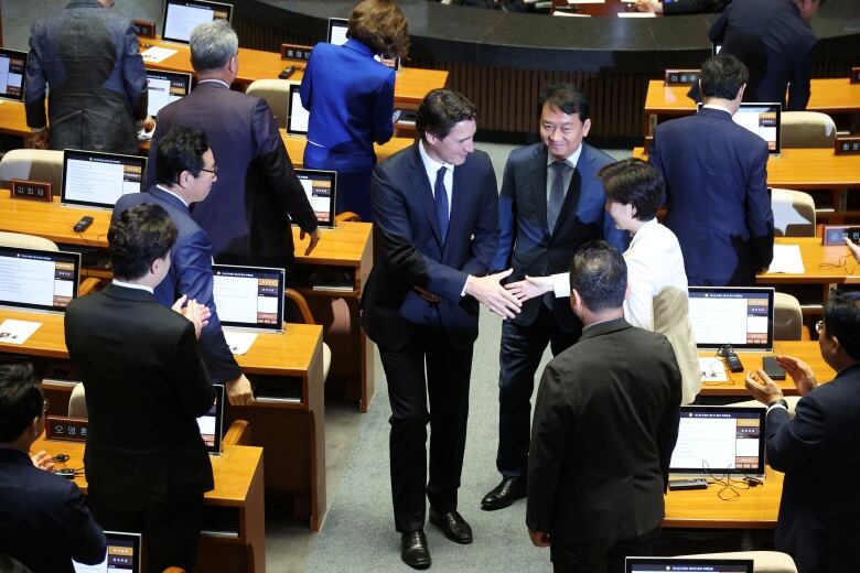 A man in a blue suit shakes hands with a woman in a white suit in a legislative assembly. Some people in the assembly hall applaud.