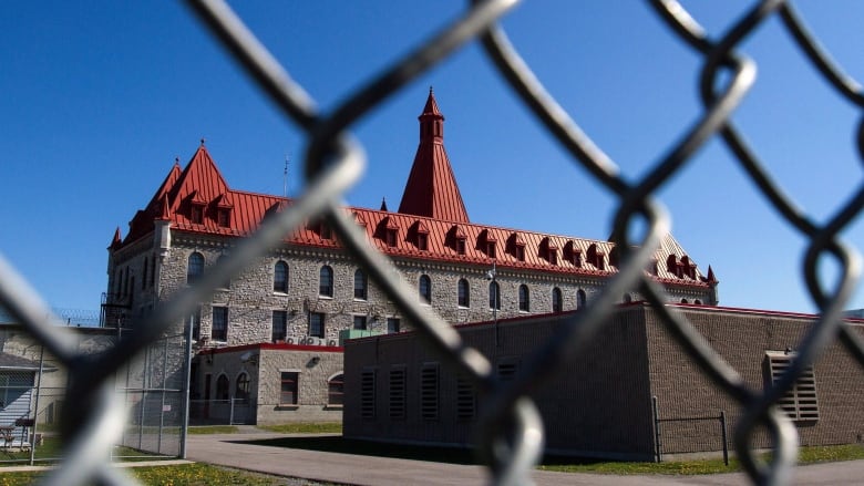 A stone building with a red roof from behind a chain link fence.