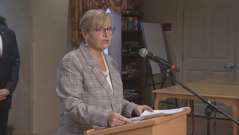 A woman with glasses and a grey checkered blazer is seen standing at a podium with a microphone in front of her.