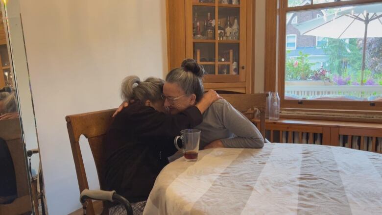 Two woman hug each other as they are seated at a table 