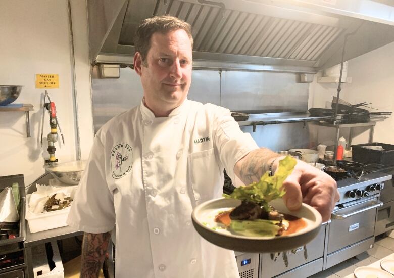 A man dressed in white standing in front of an oven holds up a plate.