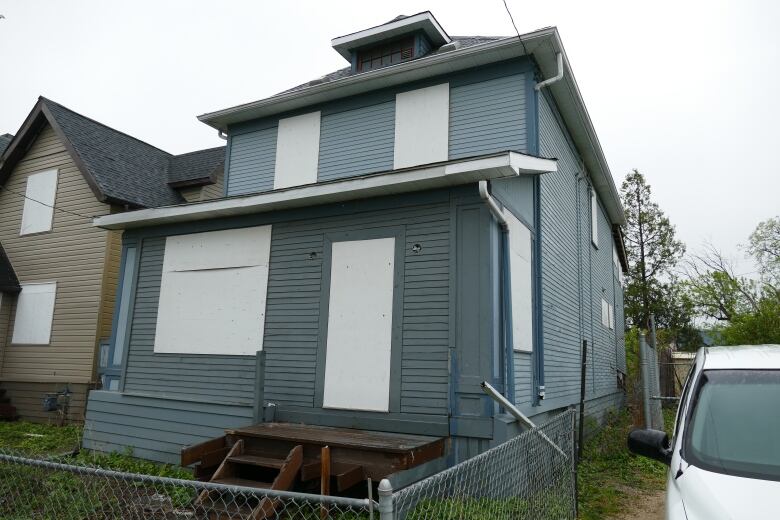 A blue two-storey house with boarded up windows. A vehicle is parked beside it.