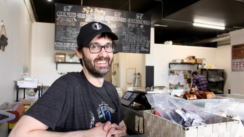 A man wearing an apron and ballcap leans on a counter, smiling. Next to him are baskets of packaged beef jerky. Hanging from the ceiling behind him is a chalkboard menu of meats for sale.