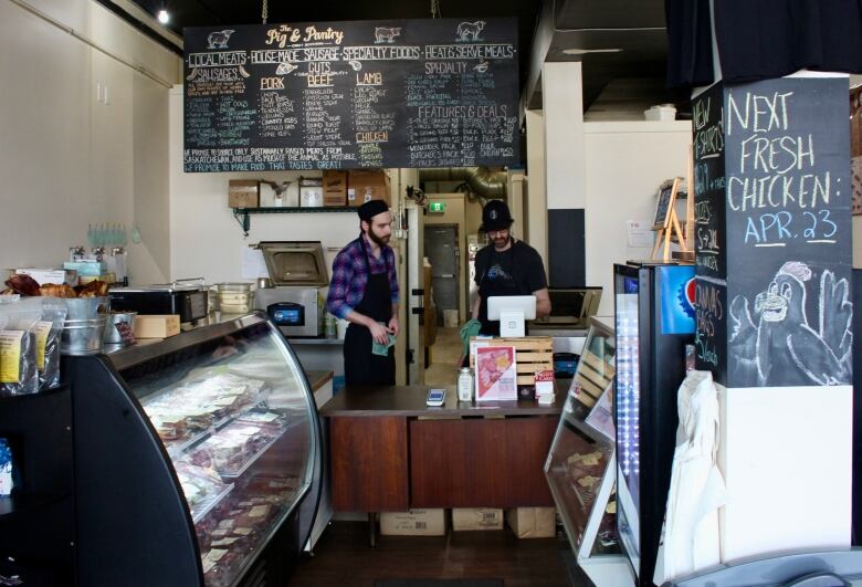 Two men wearing aprons stand behind the counter of a small butcher's shop. A chalkboard above their heads lists what's for sale, and a board to their left announces 'Next fresh chicken: Apr. 23.' A display case to their right is stocked with raw meat. 