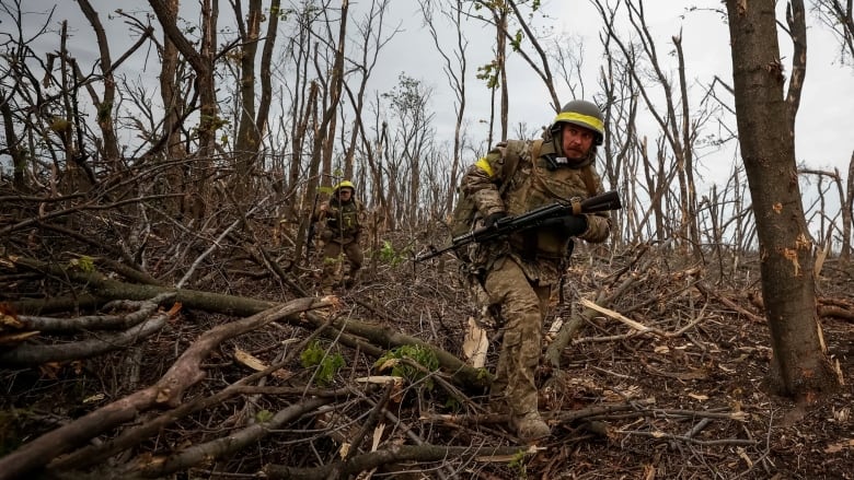 A man in military fatigues holding a weapon runs through a wooded area