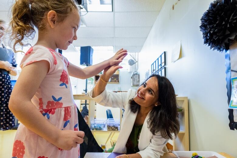 A woman in white is pictured sitting and giving a high-five to a girl standing inside a classroom.