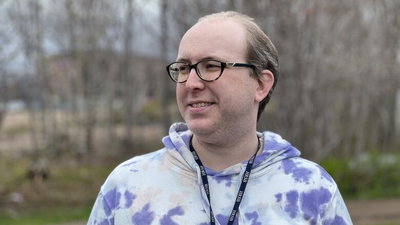 A non-binary person with glasses is wearing a hoodie and is looking to the left. They are smiling and there is greenery in the background.