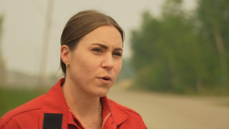 A woman wearing red speaks to a reporter by the side of a forested road.