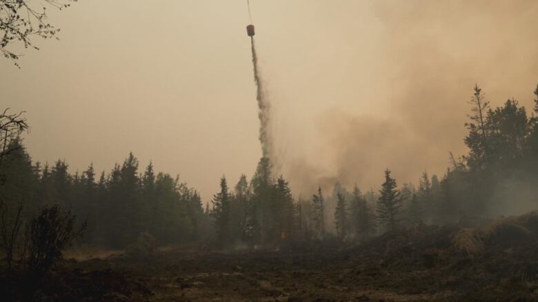 A long thin trail of fire retardant streams from an airborne bucket, presumably attached to a helicopter, into a forest full of smoke.