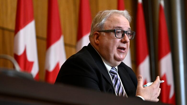 A man in a black suit, stripped tie and glasses speaks at a press conference. He is holding a pen and is sitting in front of a row of Canadian flags.