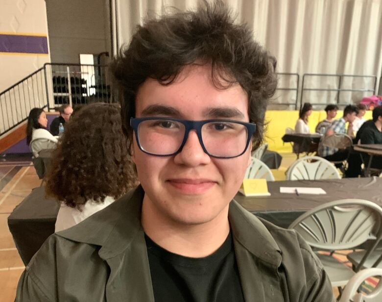 A teen boy with curly black hair and glasses smiles for the camera in a high school gymnasium with tables set up in the background.