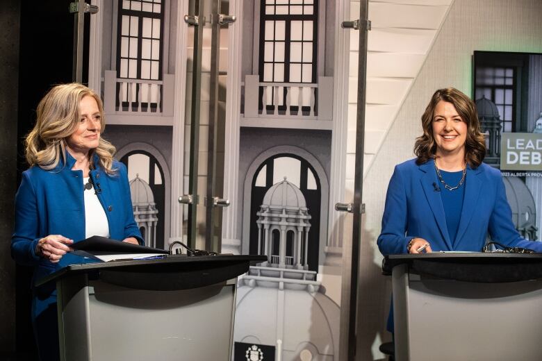 Two women wearing blue suits stand at podiums opposite each other.