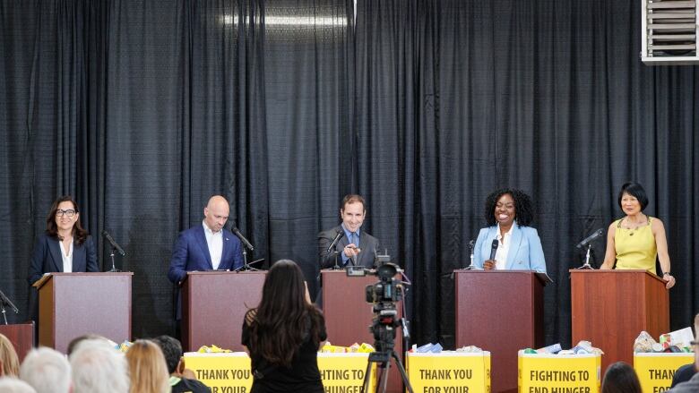 A women dressed in black takes a picture of five Toronto mayoral candidates on the debate stage. One candidate points at the photographer and smiles.