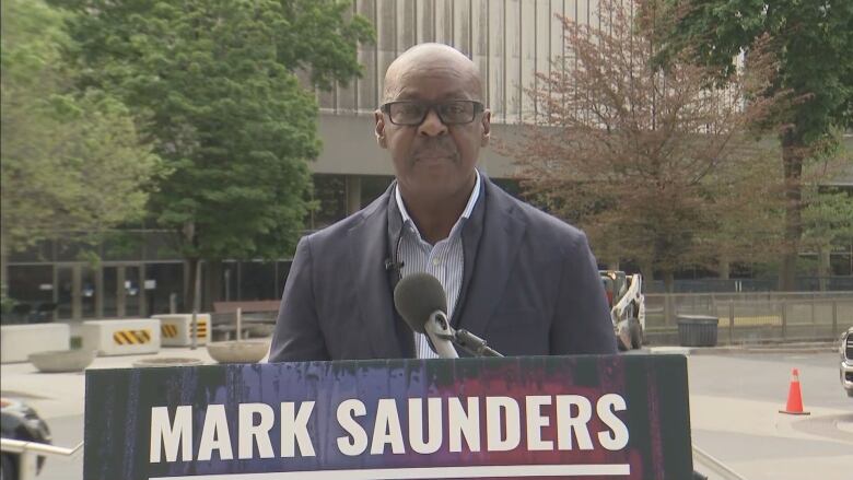 Man in a blue suit standing in front of a podium. Toronto City Hall is in the background.