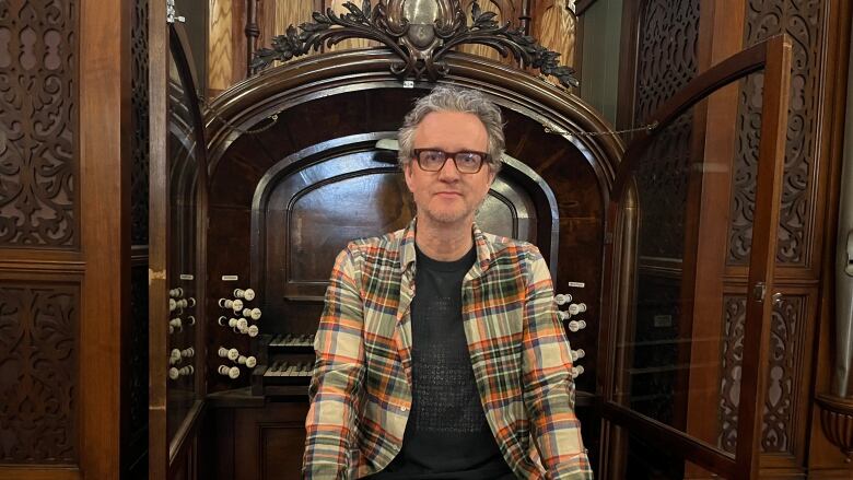 A man wearing a plaid shirt and t-shirt sits on a bench in front of an antique pipe organ. 