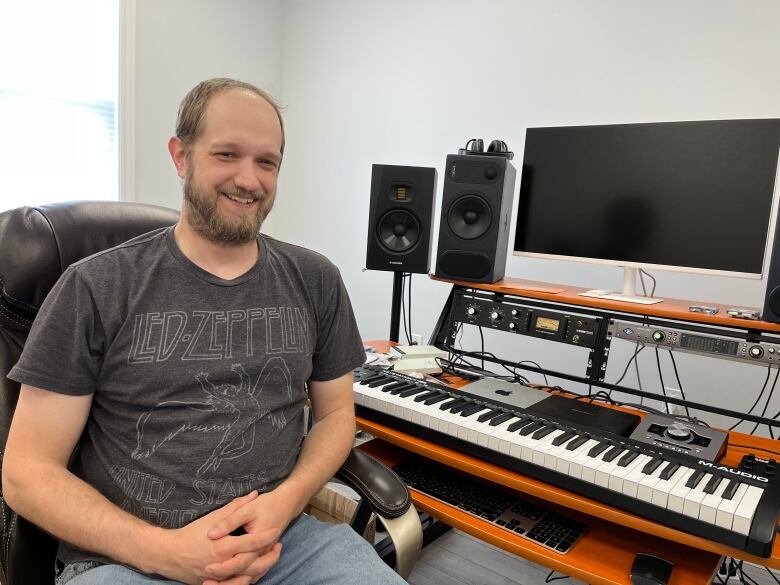 A man wearing a Led Zeppelin shirt smiles as he sits in front of a computer and recording equipment. 