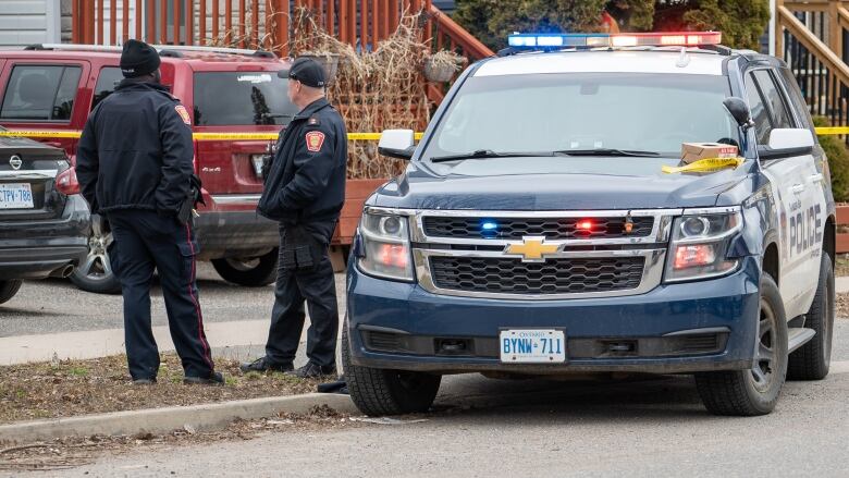 Two police officers speak with each other, they stand next to a police cruiser. 