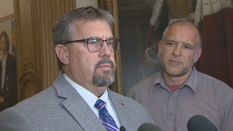 A man with short, salt and pepper hair and a goatee, wearing dark rimmed glasses, a gray suit and a blue tie with lavender diagonal stripes, looks off to the side and speaks into a few microphones. In the background, a man who looks to be in about his fifties with thinning gray hair and wearing a gray shirt with the top button undone, tilts his head listening. On the wall behind him, part of a large painting can be seen of someone wearing a royal-looking robe.