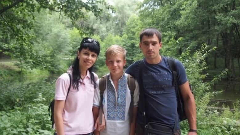 A mother and father pose with their son in a forested area.