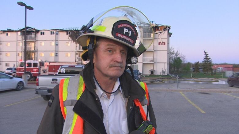 A fire chief is pictured in looking forwards. Behind him is a damaged apartment building.