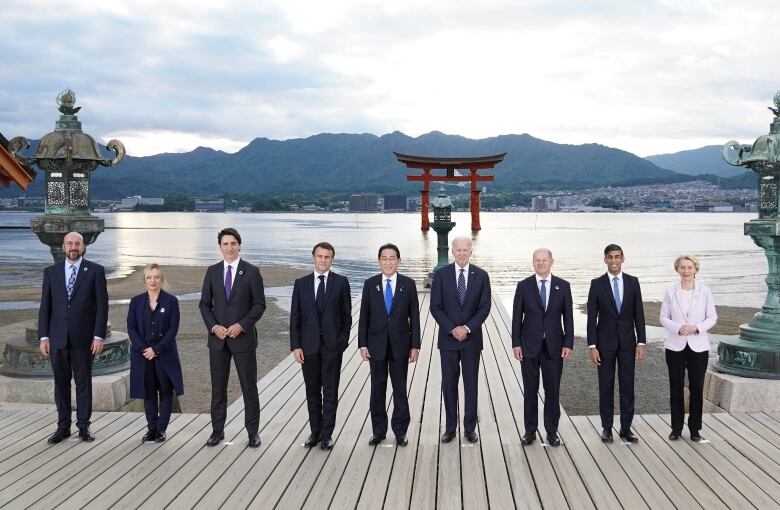 Nine people in business attire pose in a straight line for a photograph. A body of water with a torii gate can be seen behind them, as well as mountainous land beyond.