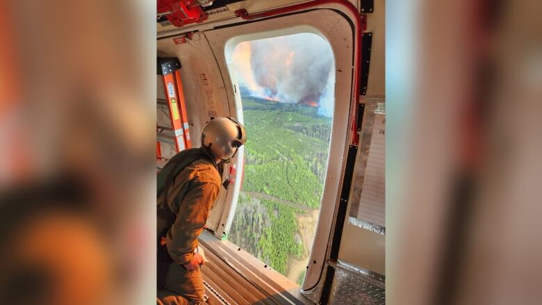 A firefighter stares out of a plane window at a large wildfire with plumes of smoke.