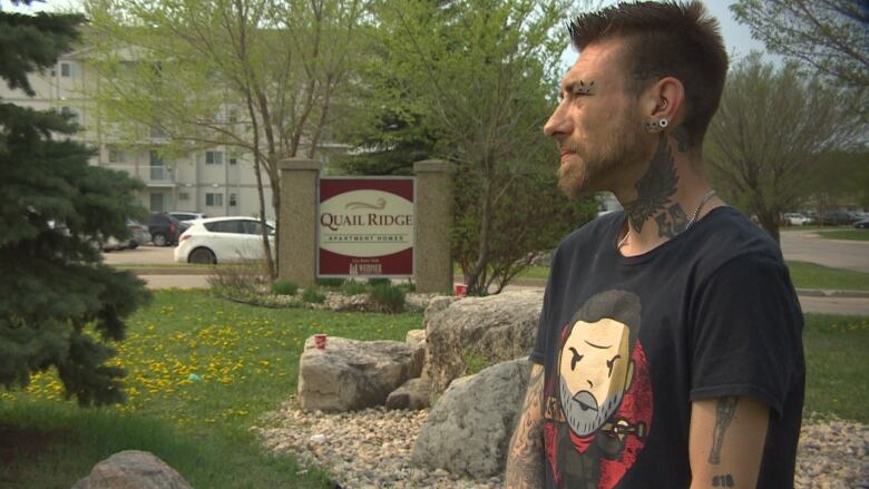 a man in a black shirt and who has tattoos stands looking off into the distance. 