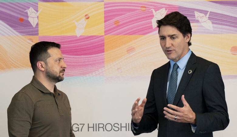 Two men, one in business attire and one bearded with a green shirt, stand near each other, seemingly talking, against a backdrop that reads 