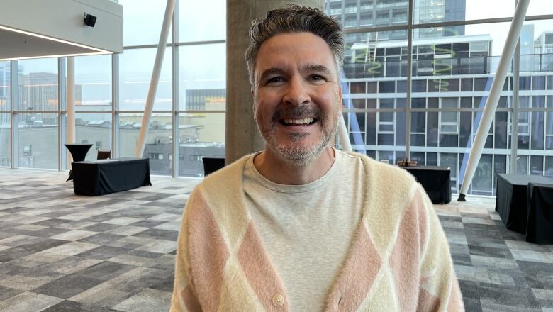 A man in a pastel argyle cardigan speaks to the camera.