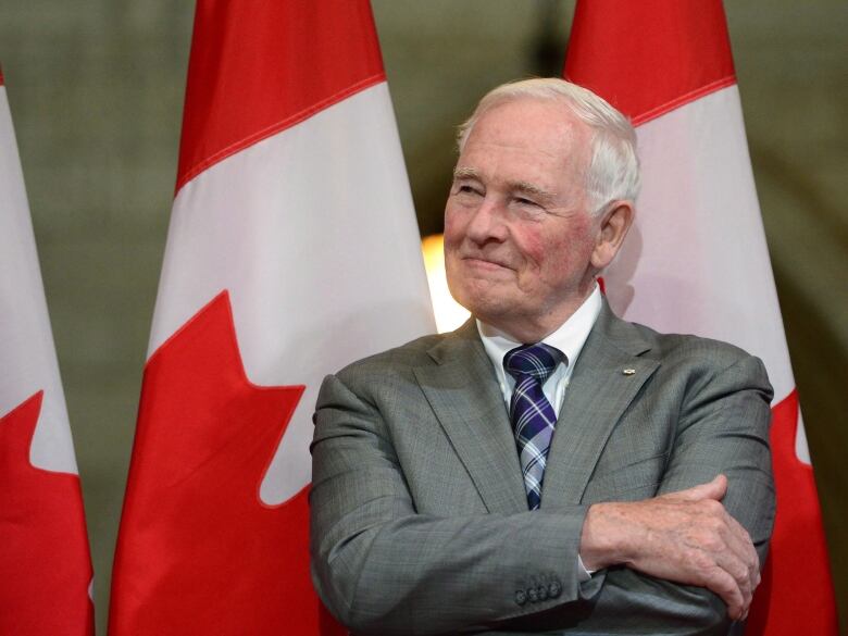 A man in a suit sits and smiles with a row of Canadian flags behind him.