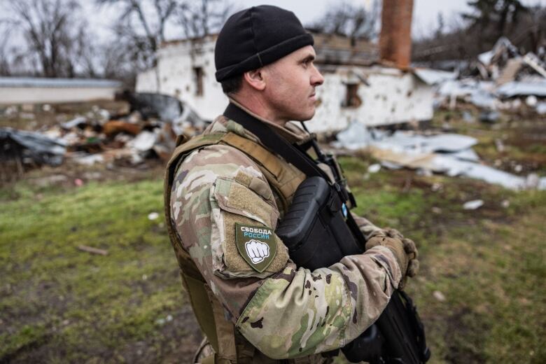 A man in military gear wearing a cap over his ears stands in profile, a dilapidated building and detritus visible behind him.