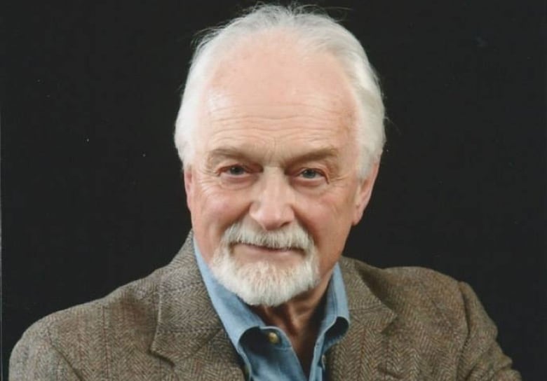 A portrait of a man with grey hair and a beard, wearing a blue collared shirt and brown tweed blazer.