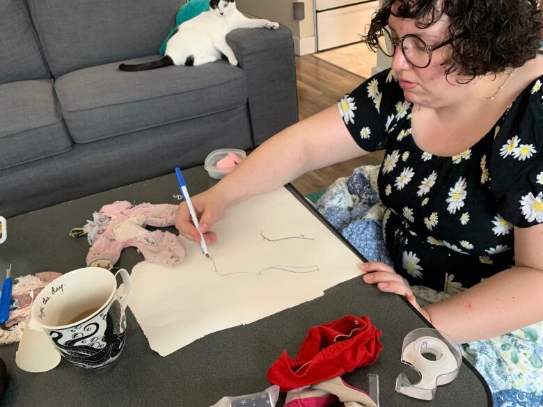 A woman draws the outline of a stuff animal on a white piece of paper. There are pieces of fabric on the table.